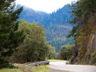 [Photo taken from a pulloff on the left side of the road looking at a curver that disappears behind a huge rock formation that extends beyond the top of the image. There are trees to the left of the guard rail on the left side and several layers of trees which extend up the mountains in the distance.]